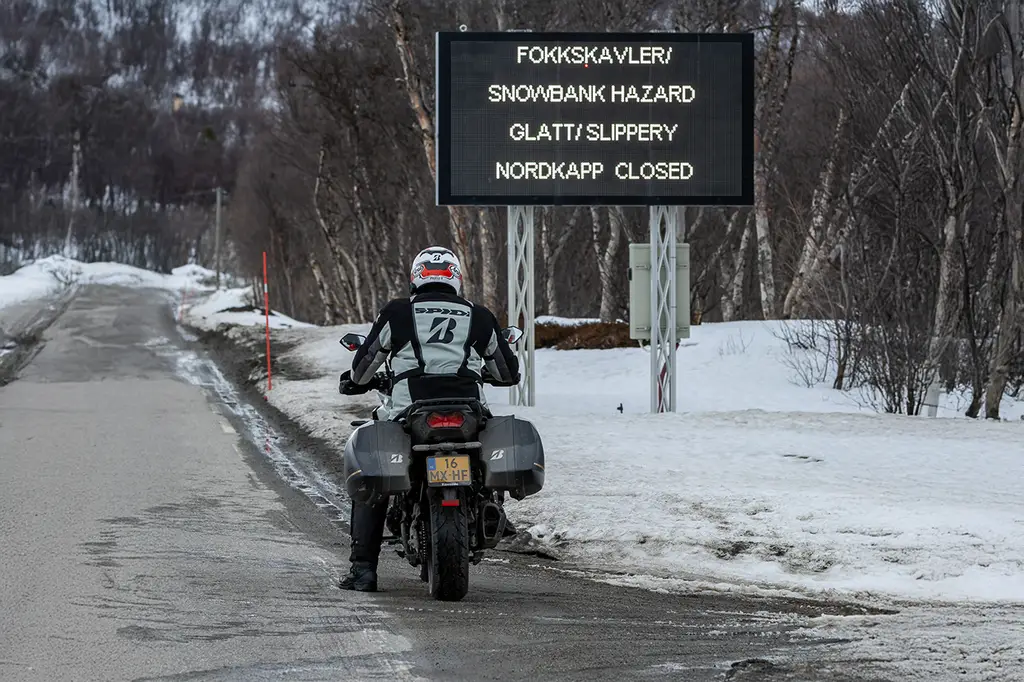 Motorcycle approaching a snowbank hazard and road closure sign near Nordkapp, highlighting icy conditions and T33 tyre reliability.