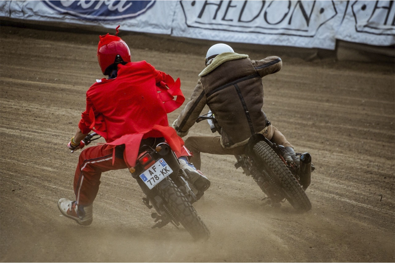 Two riders cornering side by side on vintage flat-track motorcycles, kicking up dust