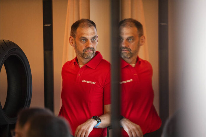 Dominique Plom, wearing a red Bridgestone polo shirt, reflected in a mirrored panel beside a motorcycle tyre.