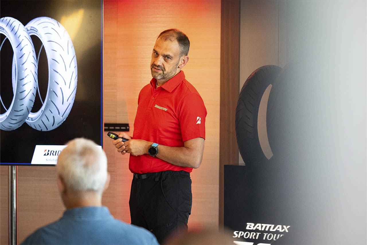 Dominique Plom presenting Bridgestone motorcycle tyres beside a screen showing tyre tread patterns.