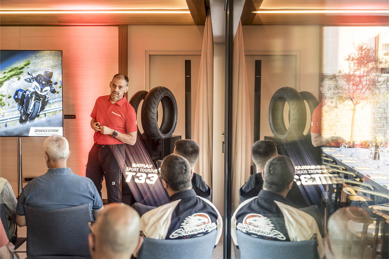 Dominique Plom presenting beside a screen showing an adventure motorcycle, with tyres on display and an audience seated.