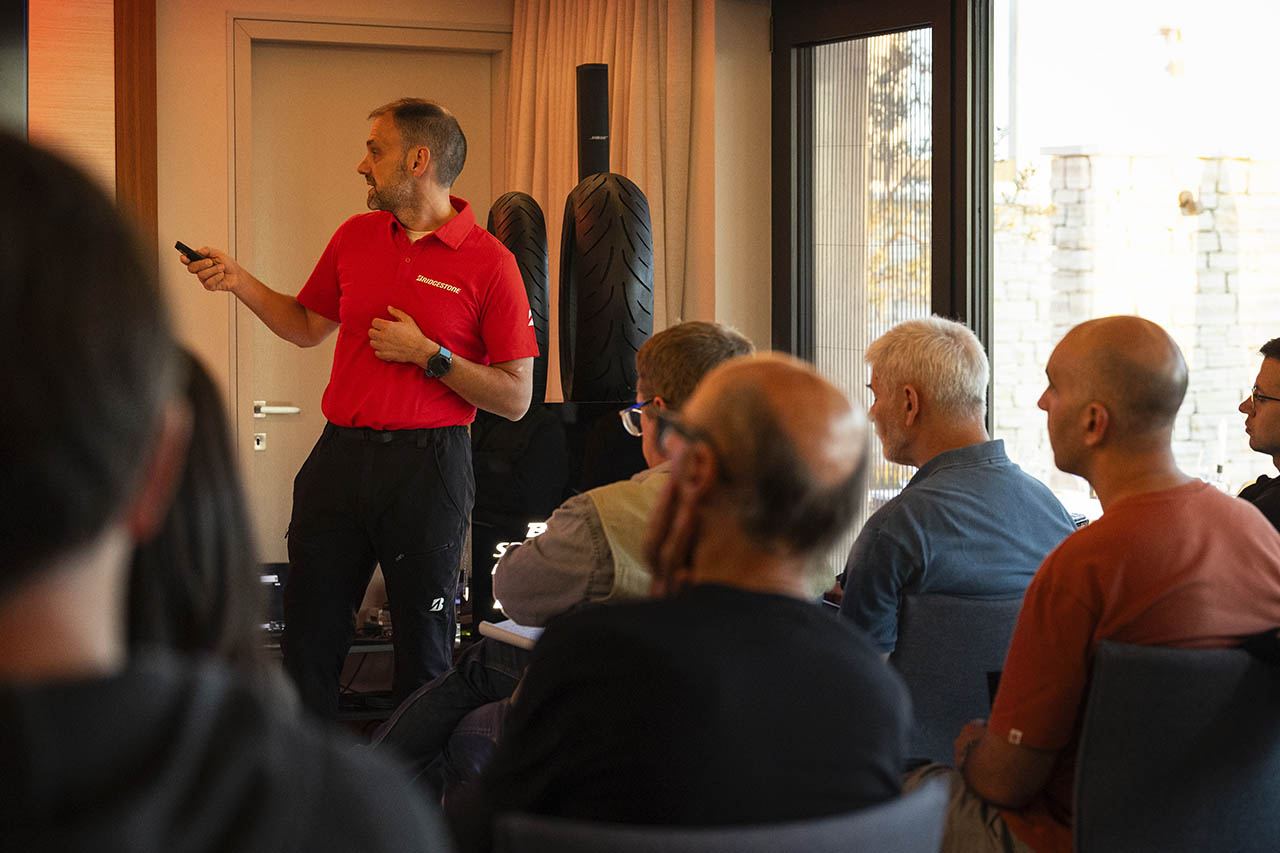 Dominique Plom presenting to a seated audience in a conference room, with motorcycle tyres displayed behind him.