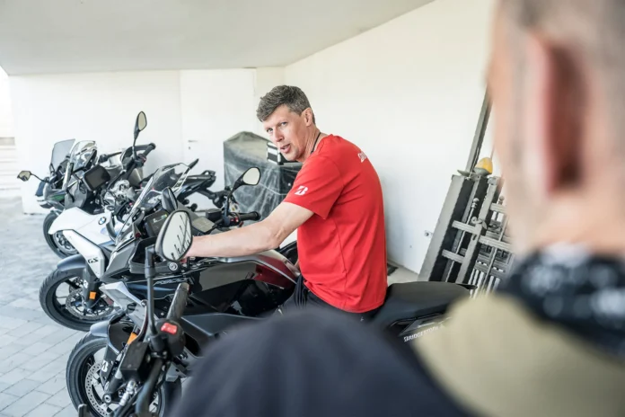 Bridgestone chief mechanic Eric sitting on a motorcycle in a garage, turning to explain something to a group of riders