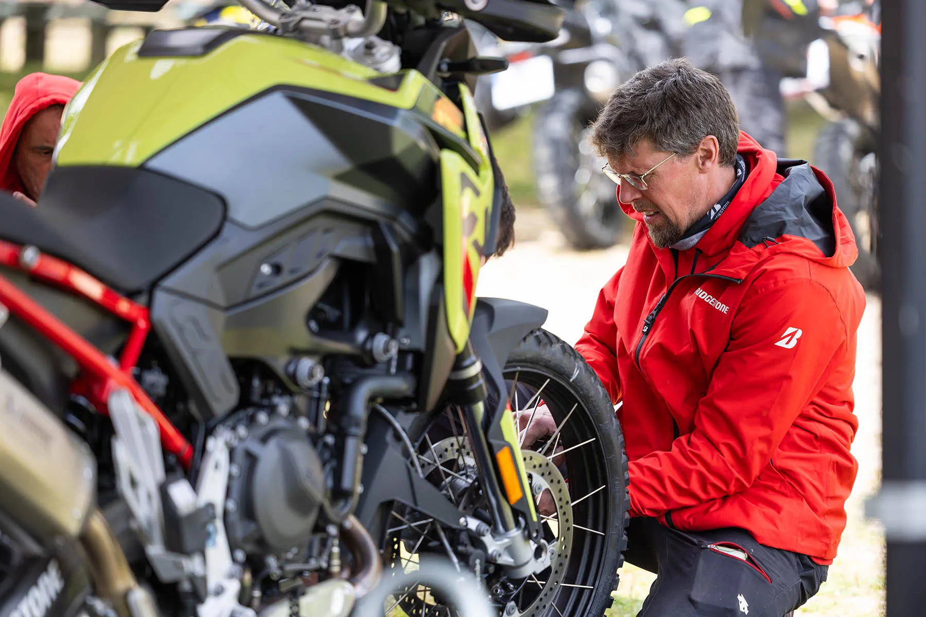 Bridgestone mechanic in a red jacket crouching beside a lime-green adventure bike, inspecting its spoked front wheel and brake disc
