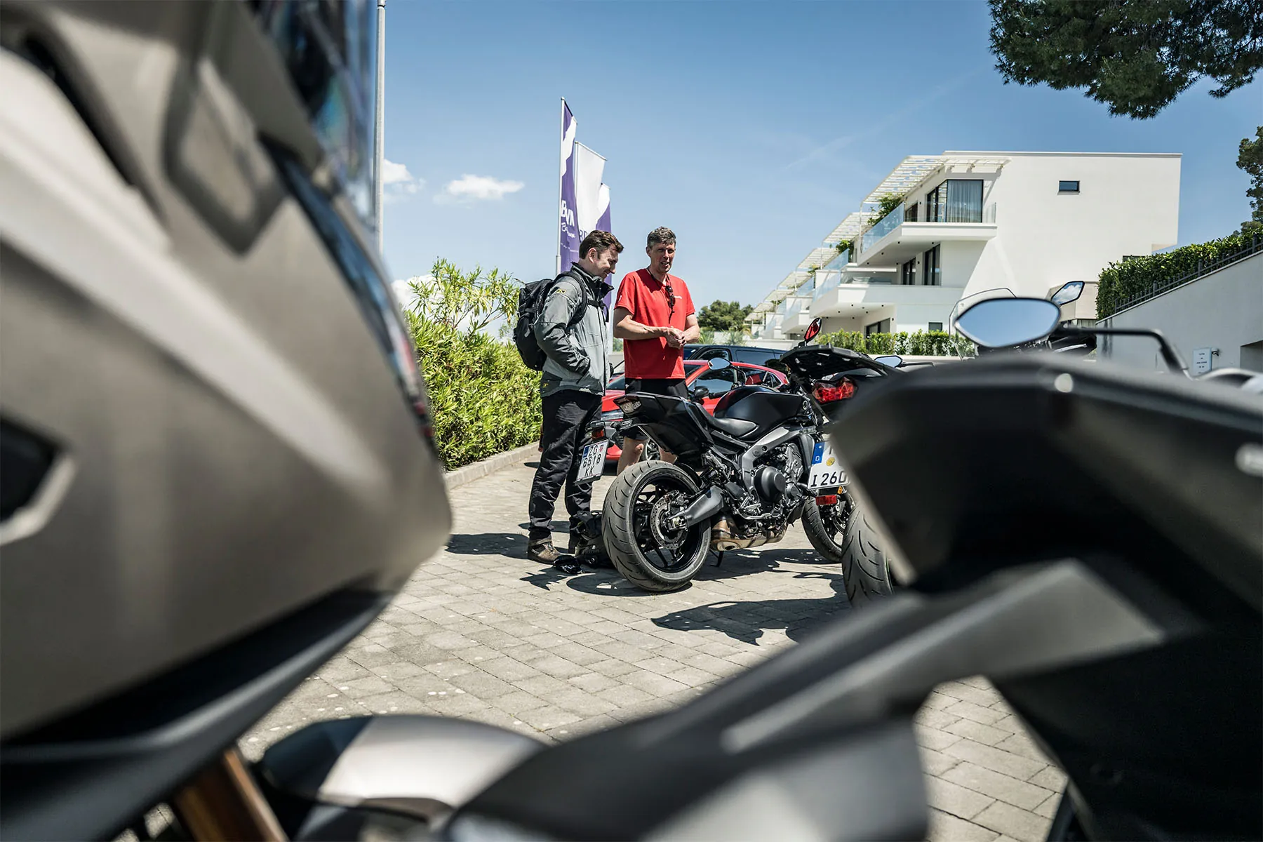 Eric in a red Bridgestone T-shirt chatting with a rider next to parked motorcycles outside a modern hotel under a blue sky
