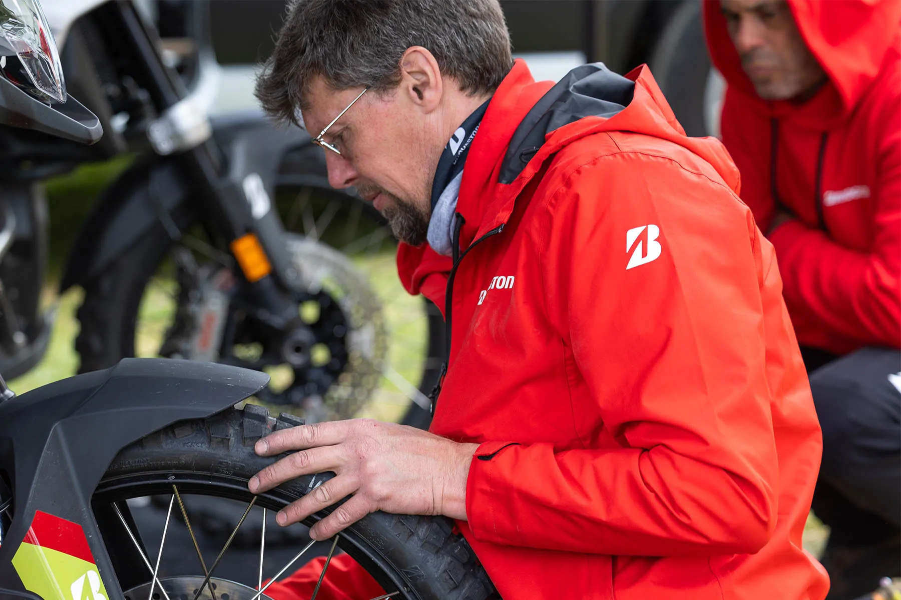 Close-up of a Bridgestone mechanic running his hand over the knobbly tread of an off-road tyre while a colleague crouches behind him