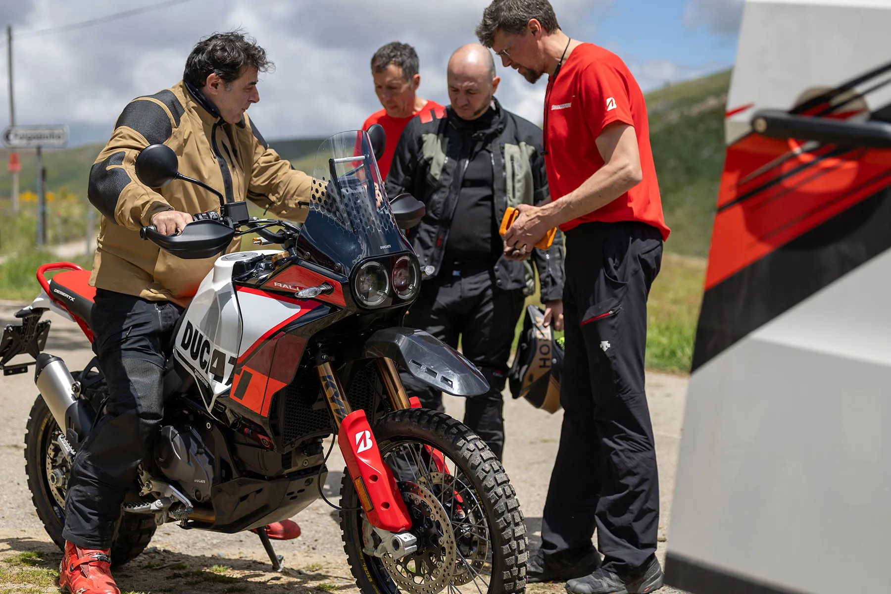Bridgestone mechanic checking a handheld device beside a Ducati DesertX Rally while three riders look on