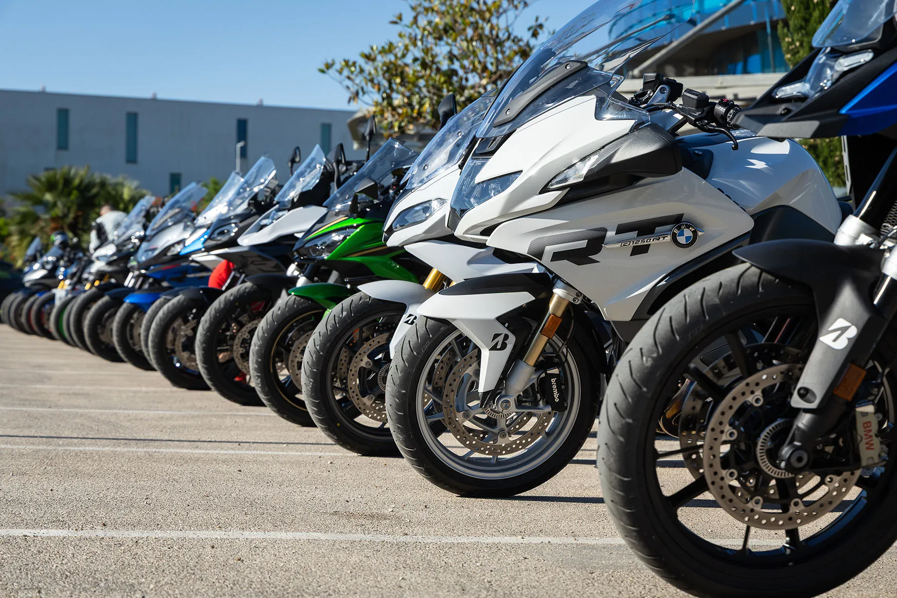 Line of parked touring and sport-touring motorcycles, including a white BMW R 1250 RT, all fitted with Bridgestone tyres in bright sunshine