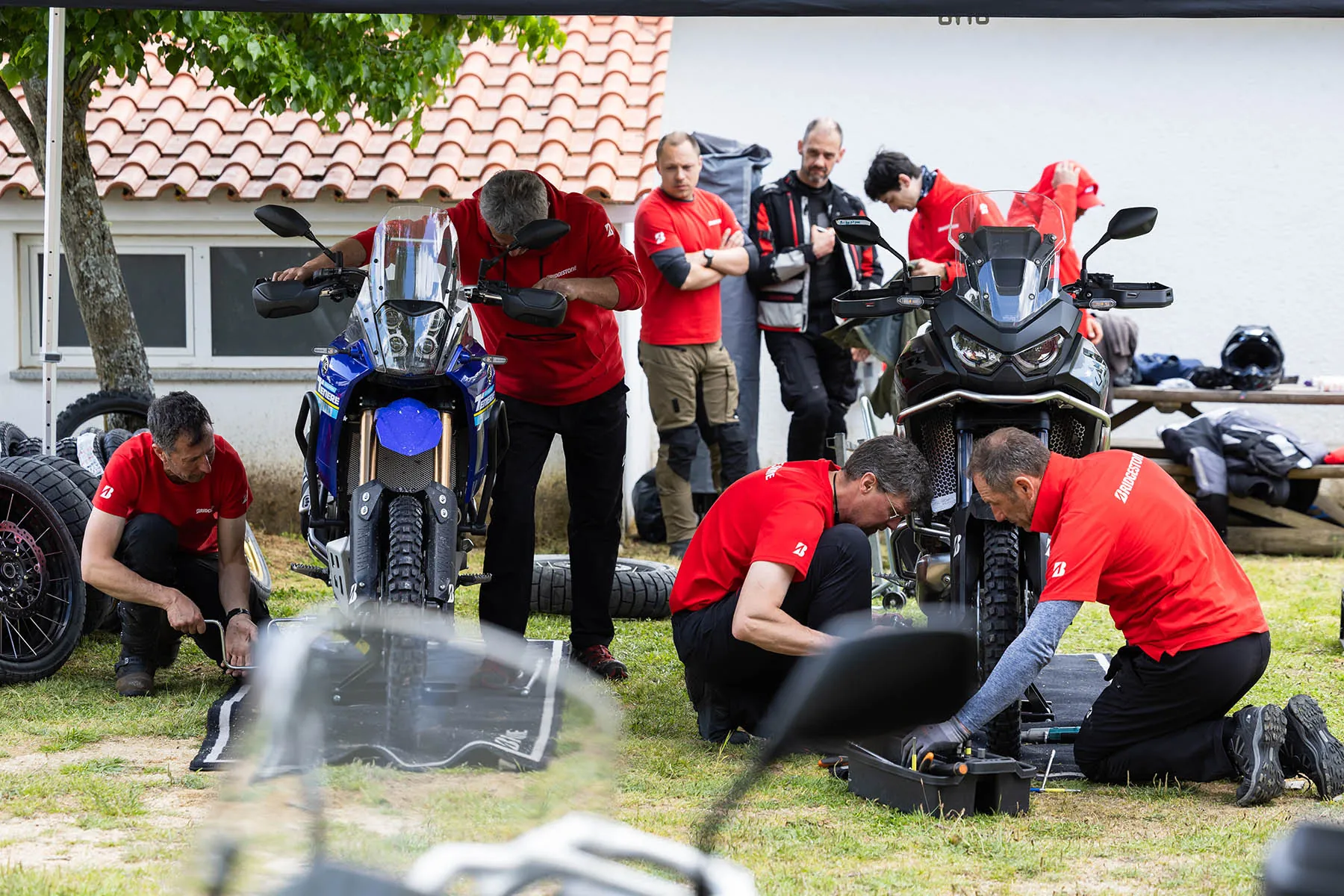 Bridgestone mechanics in red kneeling on the grass changing tyres on two adventure bikes under a tent while riders watch