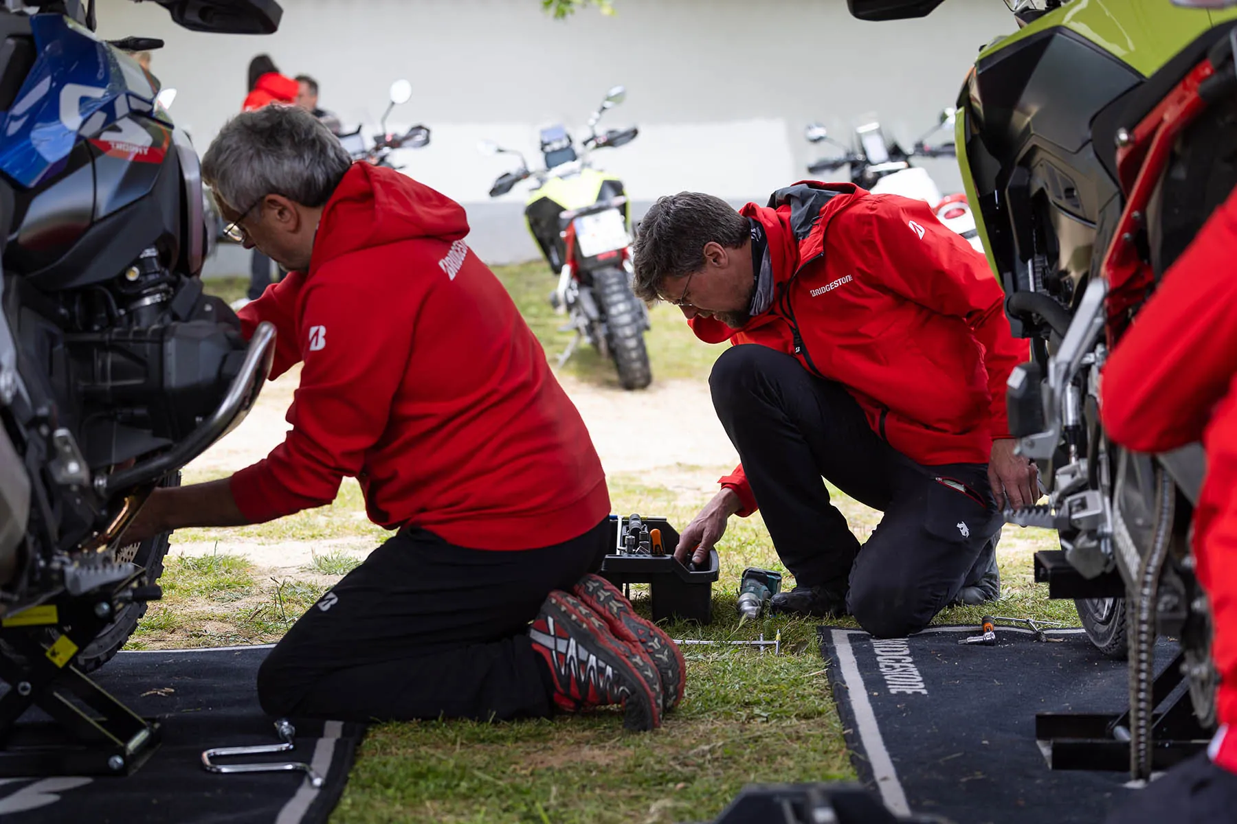Two Bridgestone mechanics kneeling on mats between adventure motorcycles, working on the wheels with a toolbox and cordless drill beside them