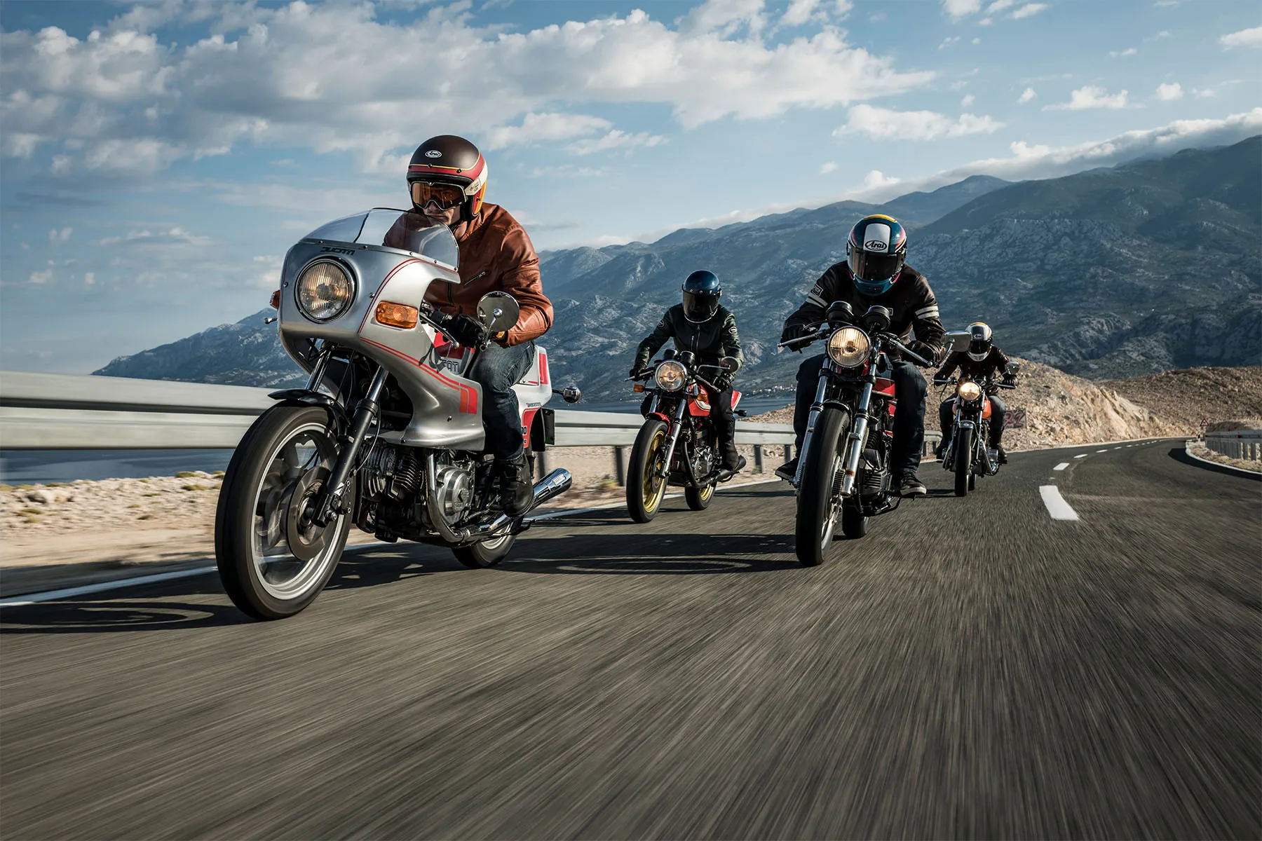 Four riders on classic motorcycles, led by a silver Ducati with a half fairing, riding along a coastal mountain road